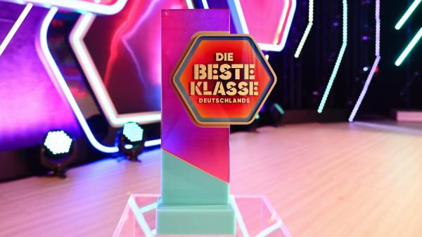 Der Pokal als hochkantige Form in pink und türkis mit dem Logo "Die beste Klasse Deutschlands" vor einer pink dunkelblauen Studiokulisse mit weiß leuchtenden Strichen und weiß umrandeten Sechsecken.