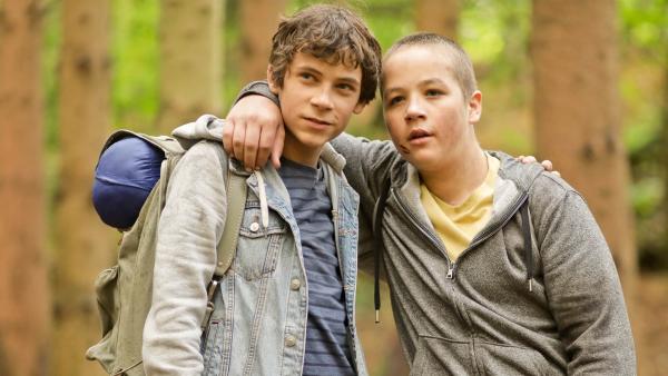 Paul und Max stehen nebeneinander im Wald. Sie haben jeweils einen Arm um die Schultern des anderen gelegt. Paul hat einen Rucksack auf den Schultern. Beide schauen erleichtert auf etwas in der Ferne.