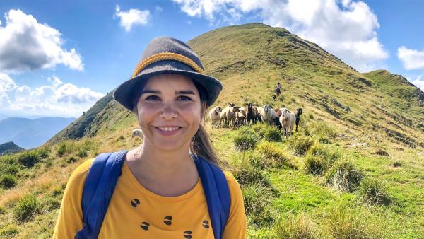 Anna lächelt. Hinter ihr Berge und eine Schafsherde.