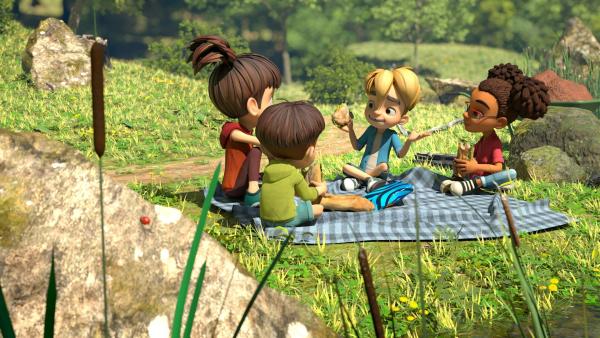 Draußen auf einer karierten Picknickdecke auf einer grünen Wiese freunden sich die Geschwister Emma und Gabriel mit zwei Kindern aus dem Dorf, Raphael und Lina, an. Alle vier Kinder sitzen im Schneidersitz auf der Decke.