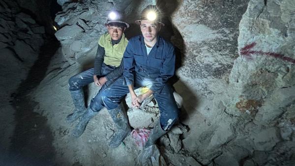 Sherif sitzt mit einem Kind in einem Bergwerk, umgeben von Steinen und Fels. Beide tragen einen Helm mit großer Stirnlampe auf dem Kopf und schmutzige Arbeitsstiefel.